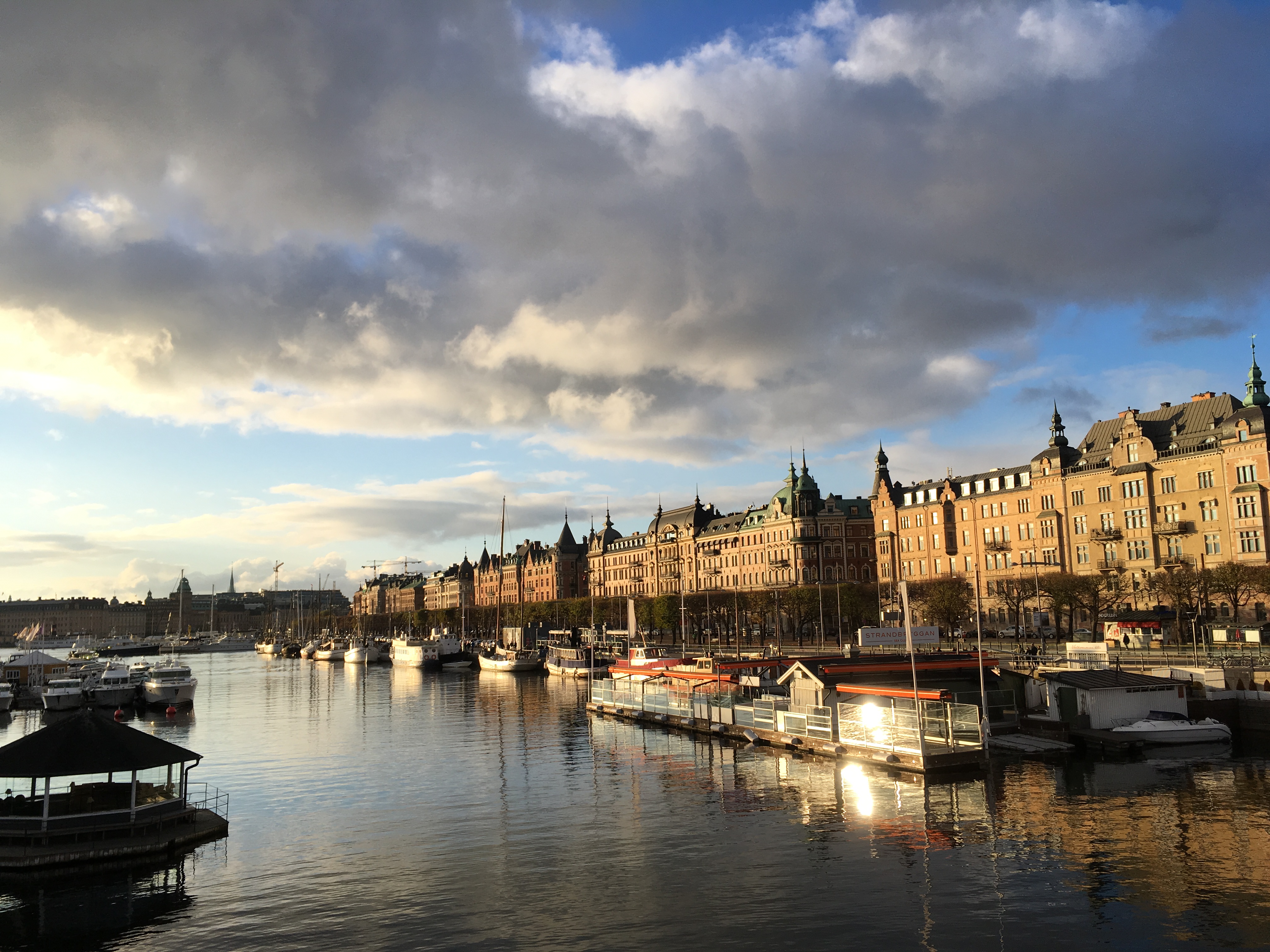 A view down a river with the afternoon sun shining blue water and gray to white clouds and a golden light on the buildings in the distance.