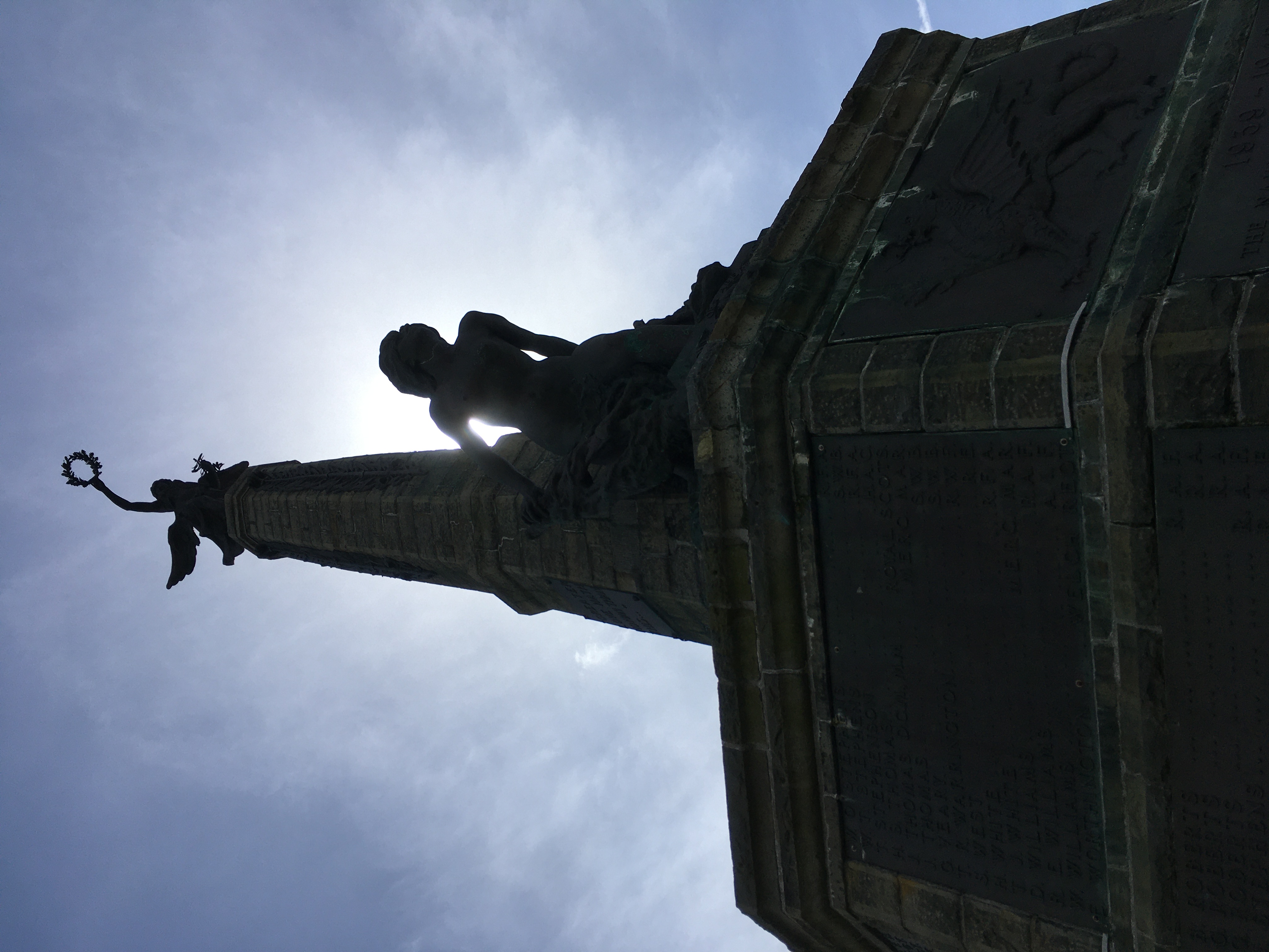 A back-lit tall monument with a wide base, a bird atop, and a figure leaning out from it. The sjy is pale blue and lightly cloudly.