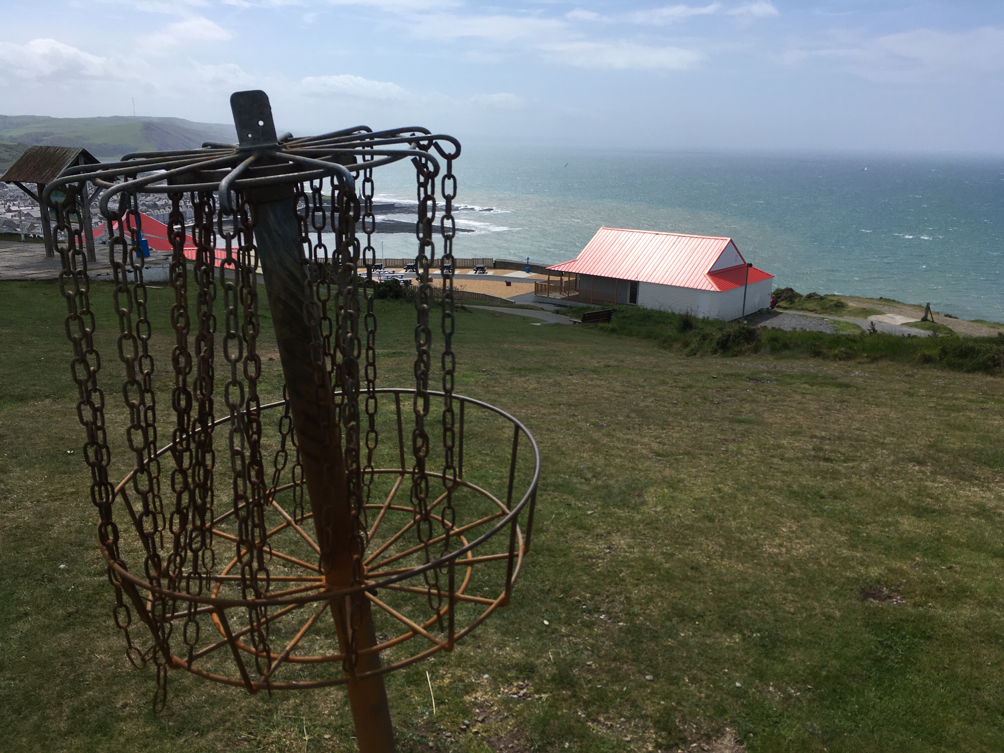 A large grassy area on a mountain overlooking some buildings and even lower, the sea. Close to the camera is a disc-golf hole with the pole and chains.