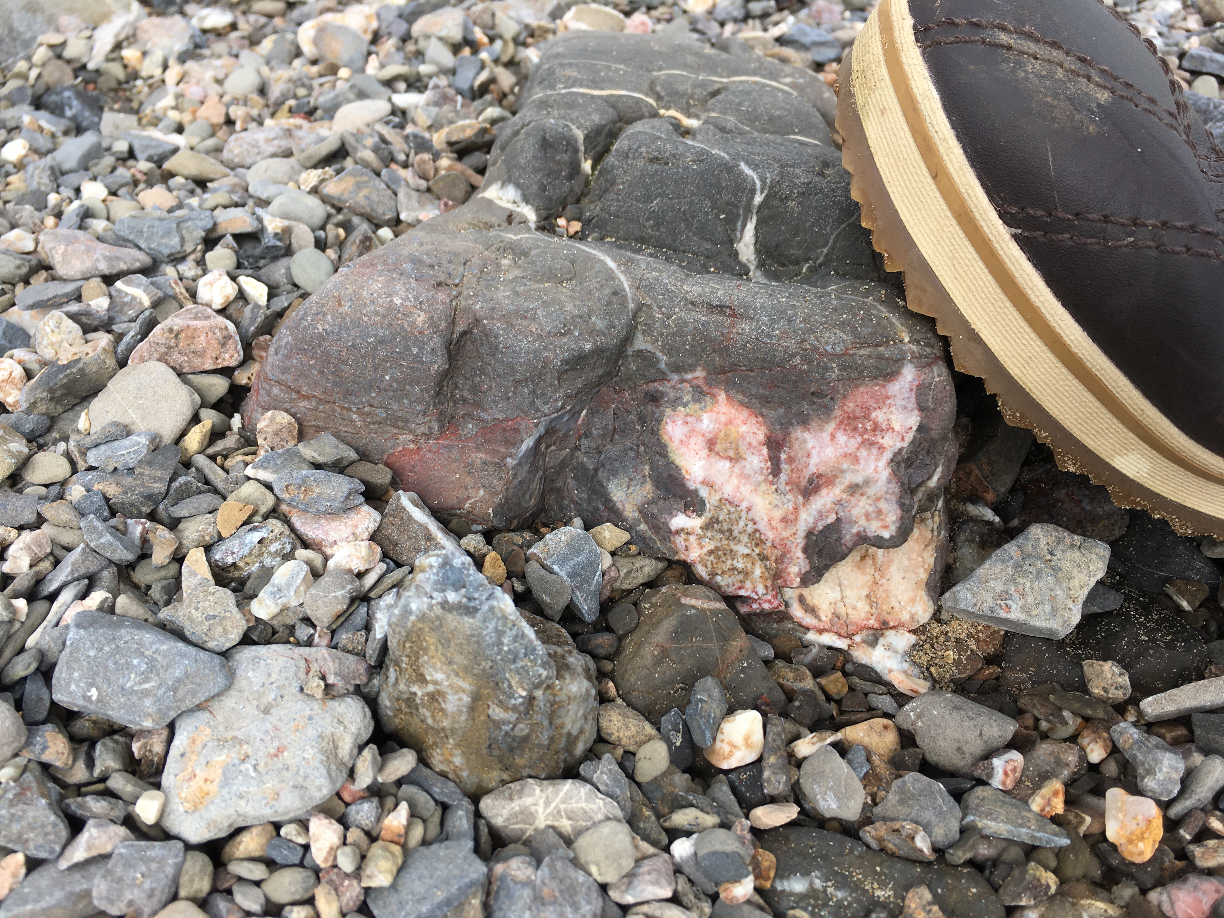 A rocky area with smaller pebbles and a larger rock with mineral formed in it looking like a pink heart. A boot rests on the big rock.