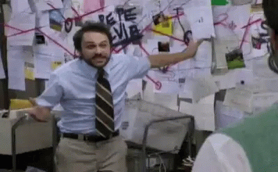 Man standing if front of a corkboard woth red strings on it flailing his arms around